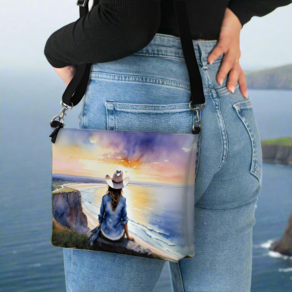 Vegan leather cross body bag imprinted with a vivid, beautiful scene of a cowgirl overlooking the ocean at sunrise. 