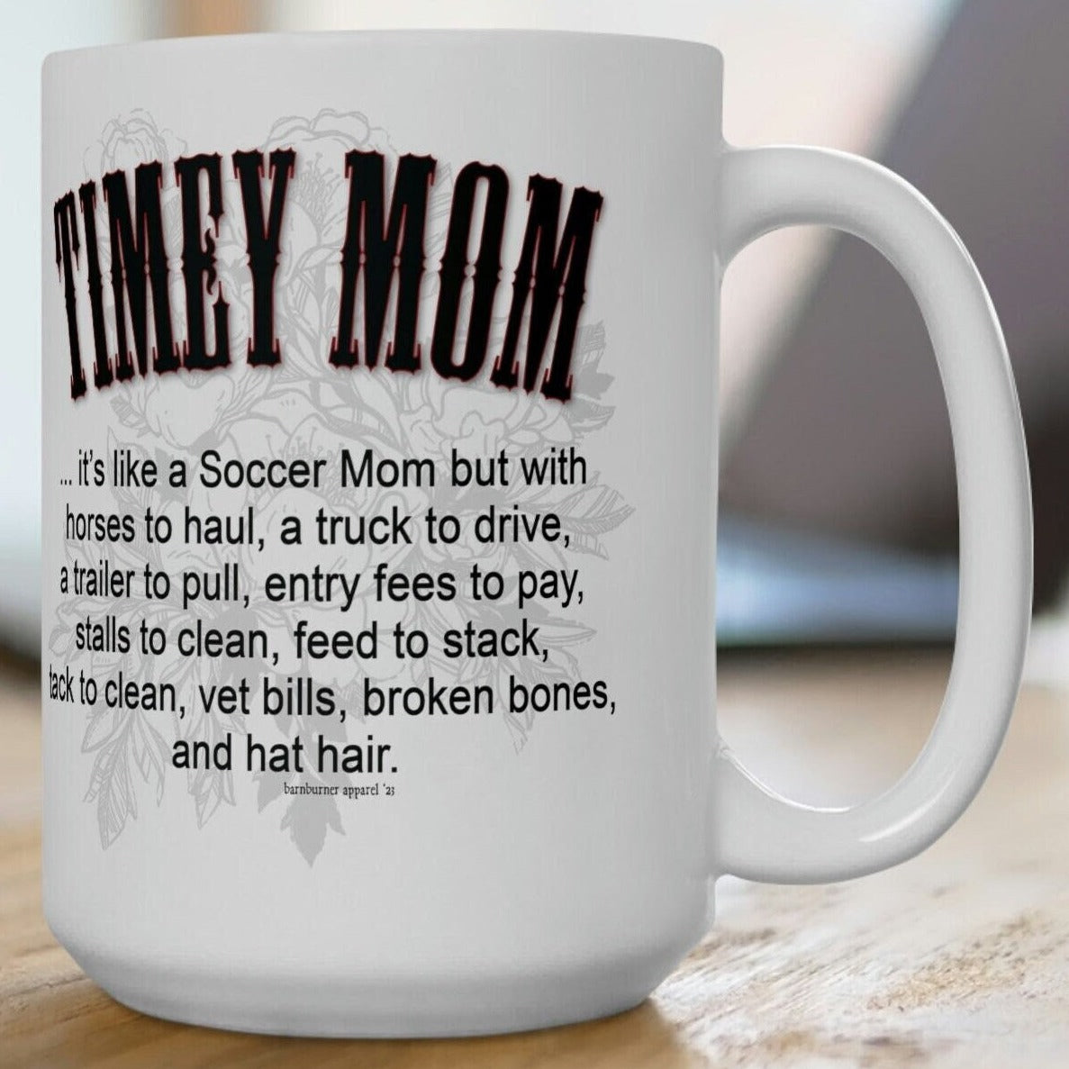 A white ceramic mug sitting on a wood picnic table. Imprint on both sides of the mug reads the same: TIMEY MOM... it's like a Soccer Mom but with horses to haul, a truck to drive, a trailer to pull, entry fees to pay, stalls to clean, feed to stack, tack to clean, vet bills, broken bones, and hat hair.