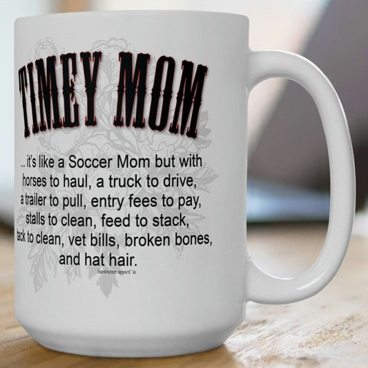 A white ceramic mug sitting on a wood picnic table. Imprint on both sides of the mug reads the same: TIMEY MOM... it's like a Soccer Mom but with horses to haul, a truck to drive, a trailer to pull, entry fees to pay, stalls to clean, feed to stack, tack to clean, vet bills, broken bones, and hat hair.