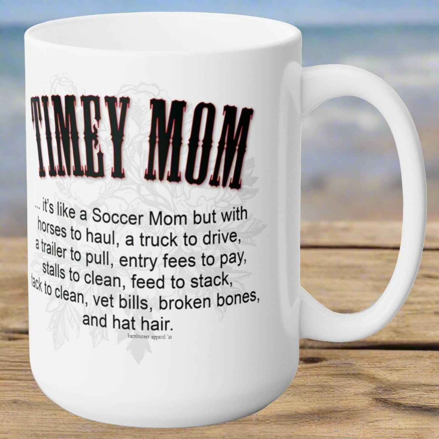 A white ceramic mug sitting on a wood picnic table. Imprint on both sides of the mug reads the same: TIMEY MOM... it's like a Soccer Mom but with horses to haul, a truck to drive, a trailer to pull, entry fees to pay, stalls to clean, feed to stack, tack to clean, vet bills, broken bones, and hat hair. 